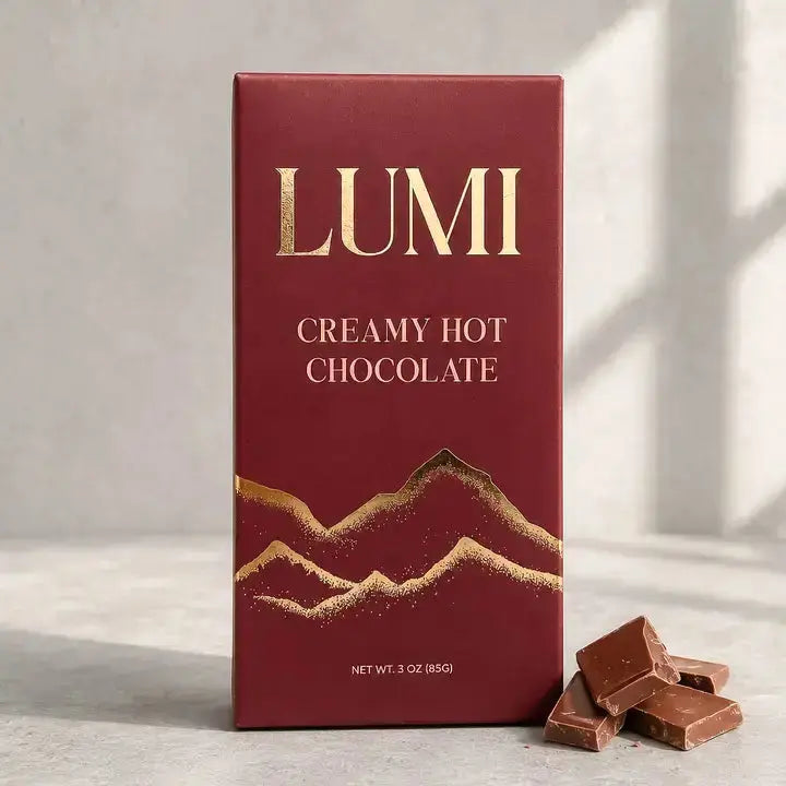 A bar of LUMI Creamy Hot Chocolate in a deep burgundy box with gold foil lettering and mountain graphics, shown standing on a light surface with a few chocolate pieces beside it.