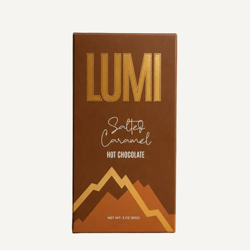 A 3 oz box of Lumi Salty Caramel Hot Chocolate, featuring a rich brown packaging design with metallic gold lettering and mountain graphics, set against a textured gray background with soft window shadows.