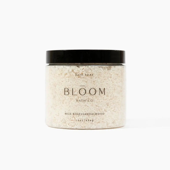 A jar of Bloom Bath + Co Wild Rose and Sandalwood bath soak, featuring a clear container filled with textured off-white salt  and a matte black lid, set against a plain white background.