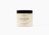 A jar of Bloom Bath + Co Wild Rose and Sandalwood bath soak, featuring a clear container filled with textured off-white salt  and a matte black lid, set against a plain white background.