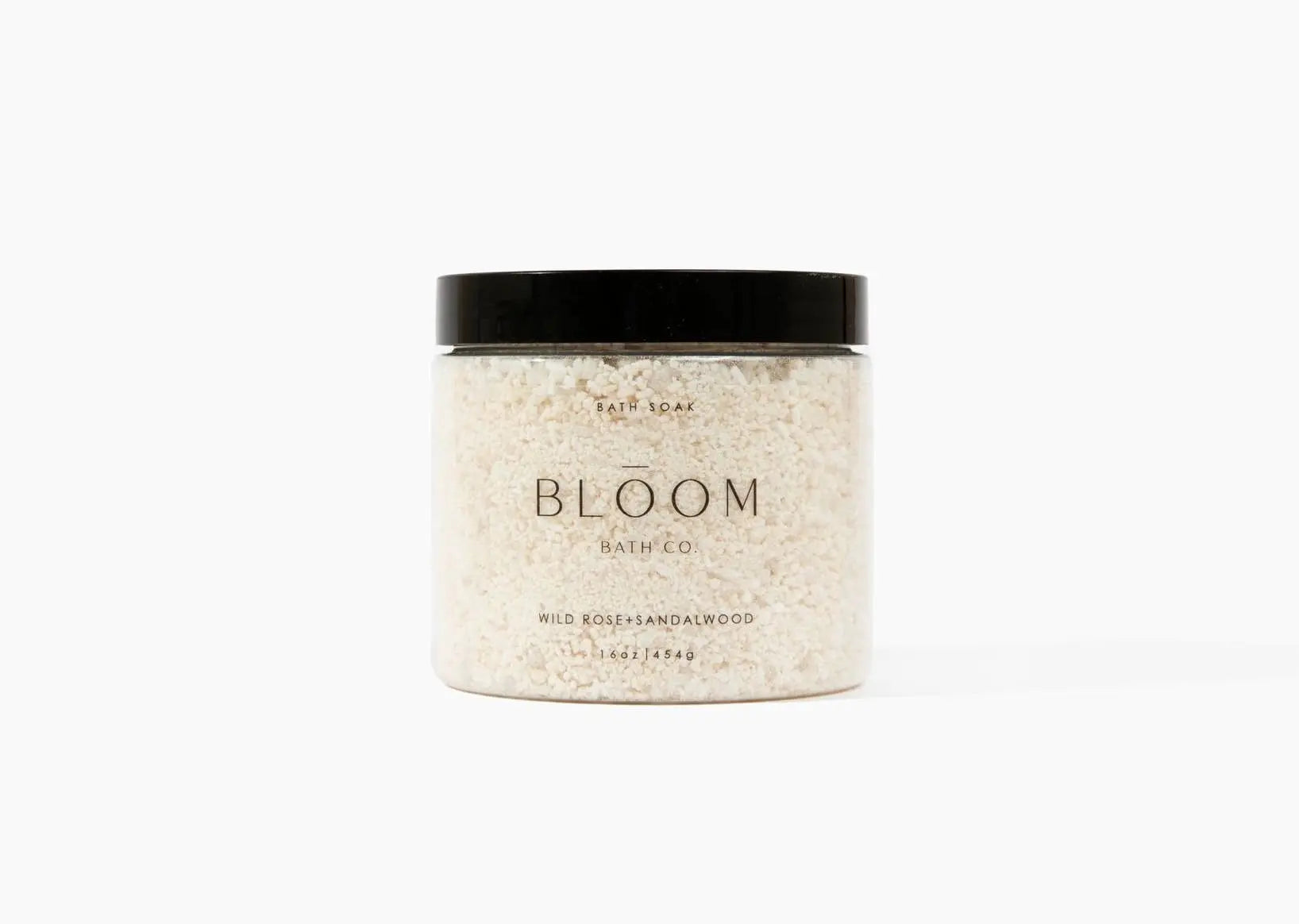 A jar of Bloom Bath + Co Wild Rose and Sandalwood bath soak, featuring a clear container filled with textured off-white salt  and a matte black lid, set against a plain white background.