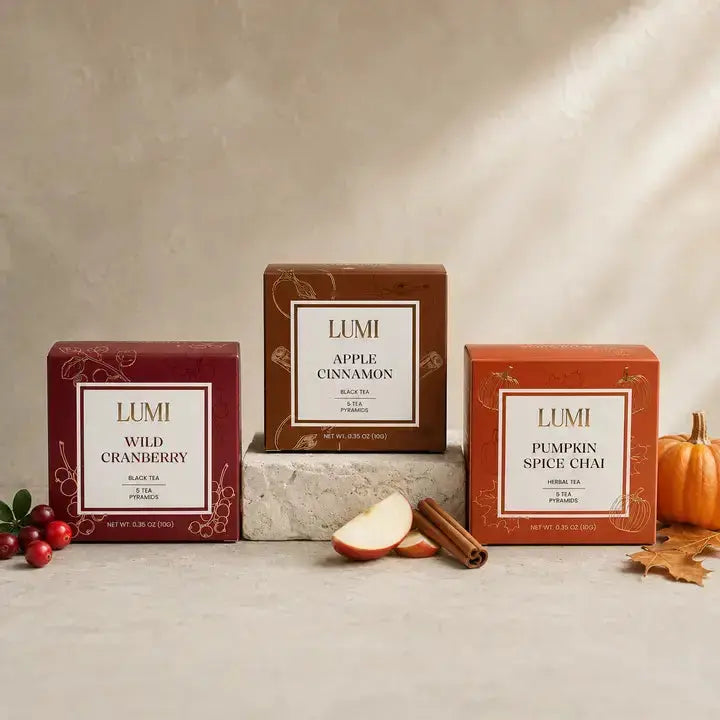 Three LUMI tea boxes arranged on a stone pedestal: Wild Cranberry in burgundy, Apple Cinnamon in brown, and Pumpkin Spice Chai in orange. The boxes are styled with cranberries, an apple slice, cinnamon sticks, and a small pumpkin on a light stone surface against a textured beige backdrop.