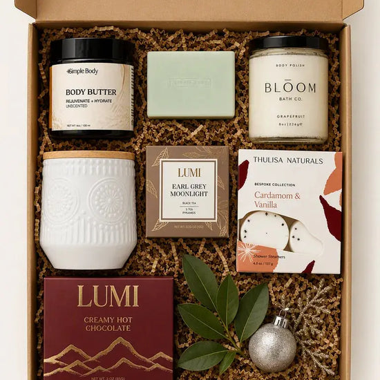 A curated gift box set containing multiple products from brands like Body Butter, Lumi, and Thuisa Naturals, arranged in a cardboard box with brown crinkle paper filler, decorated with green leaves and a silver ornament.