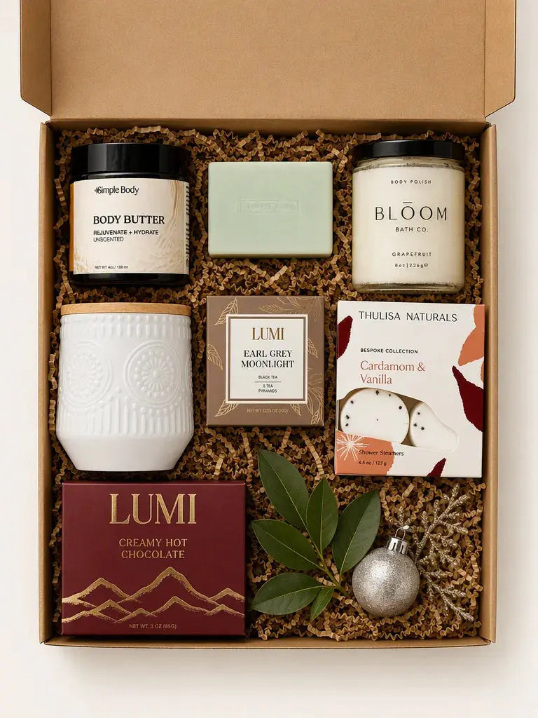 A curated gift box set containing multiple products from brands like Body Butter, Lumi, and Thuisa Naturals, arranged in a cardboard box with brown crinkle paper filler, decorated with green leaves and a silver ornament.