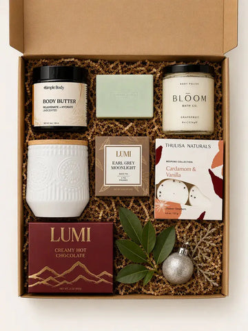 A curated gift box set containing multiple products from brands like Body Butter, Lumi, and Thuisa Naturals, arranged in a cardboard box with brown crinkle paper filler, decorated with green leaves and a silver ornament.