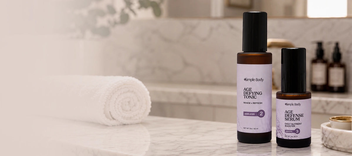 Simple Body Ageless Duo featuring Age Defying Tonic and Age Defense Serum displayed on a marble bathroom counter with a rolled white towel.