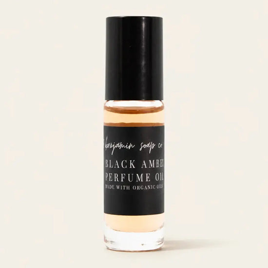 A clear glass bottle of Black Amber Perfume Oil with a black cap and black label, showing the amber-colored oil inside, set against a solid off-white background.
