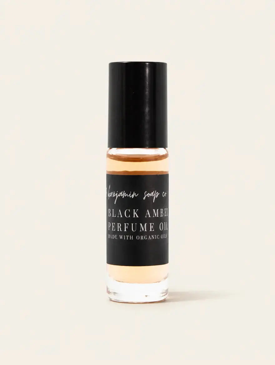 A clear glass bottle of Black Amber Perfume Oil with a black cap and black label, showing the amber-colored oil inside, set against a solid off-white background.