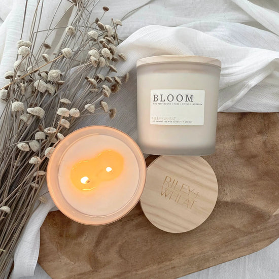 Candle labeled "Bloom" by Riley + Wheat showing it burning cleanly sitting next to dried plants on a wooden board with a linen cloth.