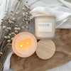 Candle labeled "Bloom" by Riley + Wheat showing it burning cleanly sitting next to dried plants on a wooden board with a linen cloth.