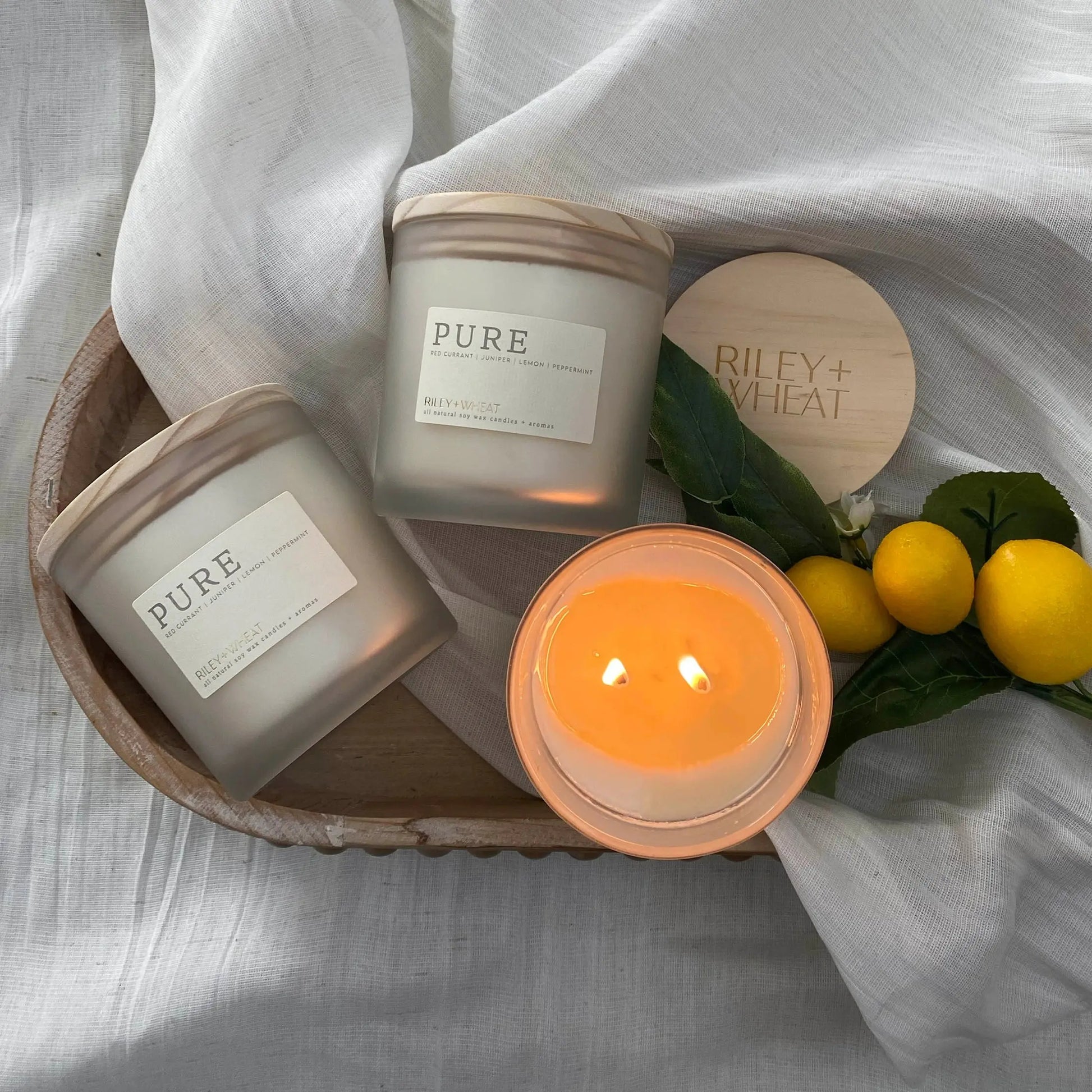 Candle labeled "PURE" by Riley + Wheat showing a double wick burning with fresh lemons next to in on a soft textured white linen background.