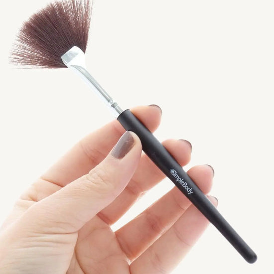 A hand holding a sleek black fan brush with synthetic bristles and a Simple Body logo printed on the handle.