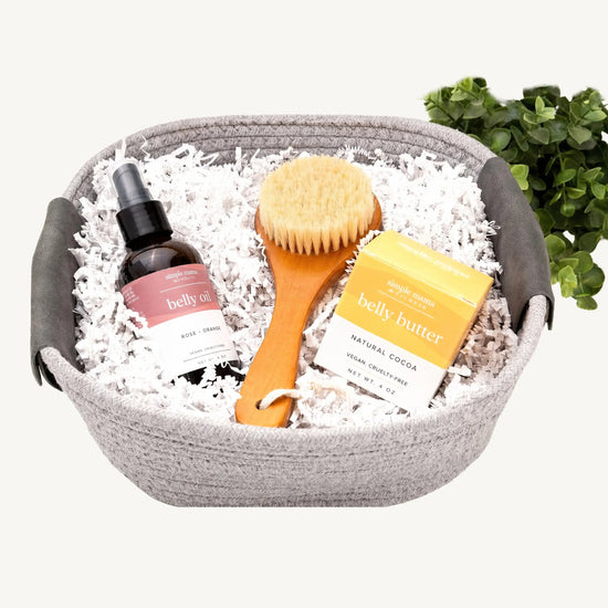 A curated wellness gift set in a gray woven basket, featuring a bottle of Simple Mama Belly Oil, a wooden body brush, and a box of Simple Mama Belly Butter nestled in white shredded paper, with a small green plant in the background.