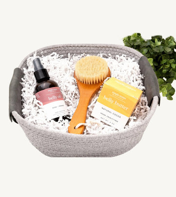 A curated wellness gift set in a gray woven basket, featuring a bottle of Simple Mama Belly Oil, a wooden body brush, and a box of Simple Mama Belly Butter nestled in white shredded paper, with a small green plant in the background.