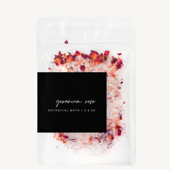 A clear stand-up pouch of botanical bath salts labeled 'geranium rose' with a black label, filled with white bath salts and topped with dried red and orange flower petals, on a light off-white background.