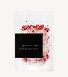 A clear stand-up pouch of botanical bath salts labeled 'geranium rose' with a black label, filled with white bath salts and topped with dried red and orange flower petals, on a light off-white background.