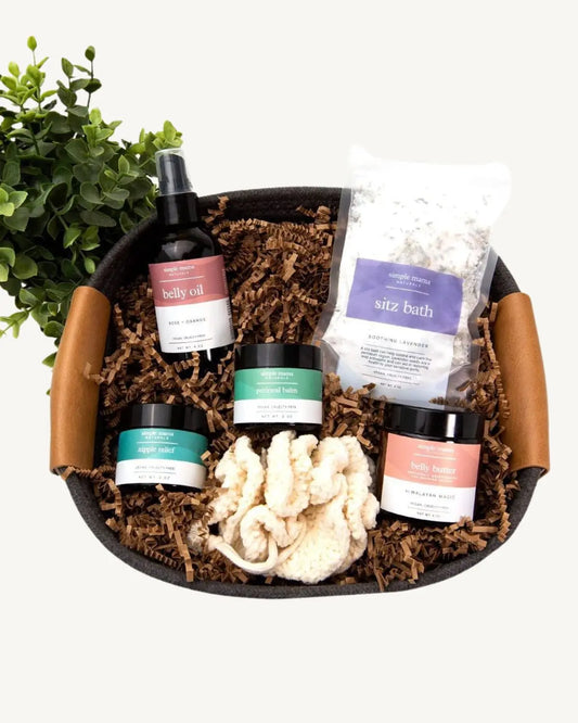 A gift basket containing various skincare products including a sitz bath packet, belly oil, belly butter, perineal and nipple balms, and a cotton pouf, all arranged in a reusable cotton rope basket.