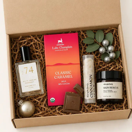 An open kraft gift box filled with spa-style products on a white background. Visible items include a Lake Champlain Chocolates Classic Caramel milk chocolate bar, a bottle labeled 74, a lip balm tube labeled Cinnamon, a jar labeled Skin Rescue, a chocolate piece, greenery with silver ornaments, and a gold sphere, all arranged in brown paper shred.