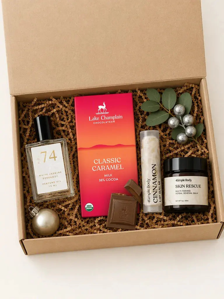 An open kraft gift box filled with spa-style products on a white background. Visible items include a Lake Champlain Chocolates Classic Caramel milk chocolate bar, a bottle labeled 74, a lip balm tube labeled Cinnamon, a jar labeled Skin Rescue, a chocolate piece, greenery with silver ornaments, and a gold sphere, all arranged in brown paper shred.