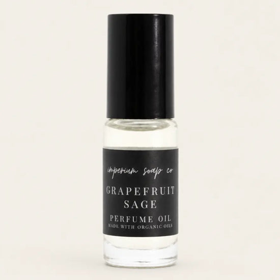 A clear glass bottle of Grapefruit Sage perfume oil by Benjamin Soap Co. with a black cap and a black label, shown against a solid light beige background. 
