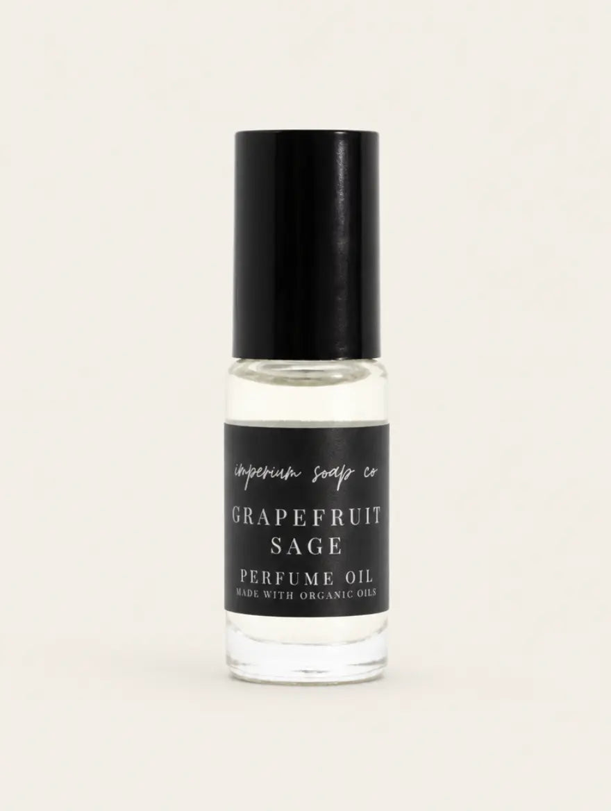 A clear glass bottle of Grapefruit Sage perfume oil by Benjamin Soap Co. with a black cap and a black label, shown against a solid light beige background. 