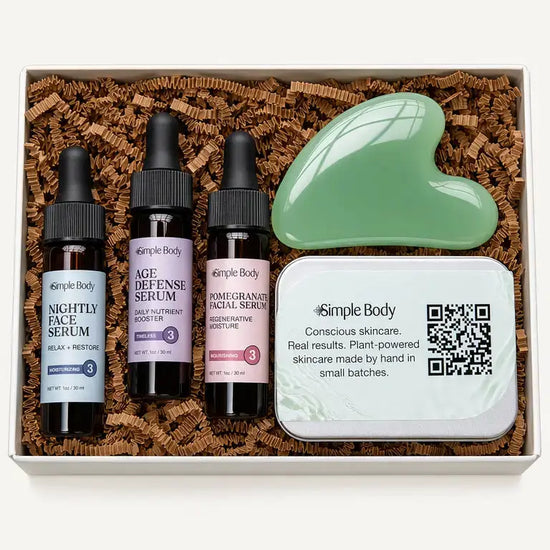 A skincare gift set from Simple Body arranged in a white box filled with brown crinkle paper. The set includes three small dropper bottles labeled Nightly Face Serum, Age Defense Serum, and Pomegranate Facial Serum, a mint green gua sha face scraper, and a white branded tin.