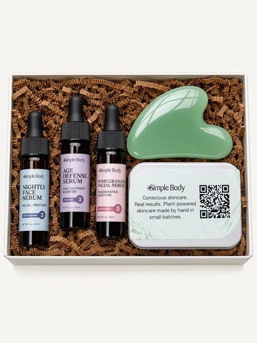 A skincare gift set from Simple Body arranged in a white box filled with brown crinkle paper. The set includes three small dropper bottles labeled Nightly Face Serum, Age Defense Serum, and Pomegranate Facial Serum, a mint green gua sha face scraper, and a white branded tin.