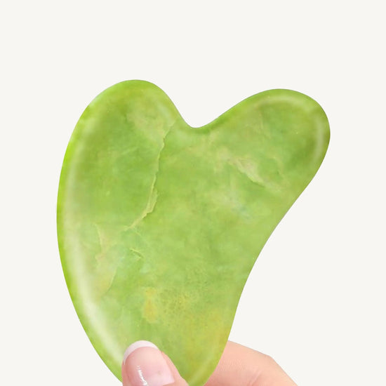 Two fingers holding a green Jade Gua Sha tool on a white background.
