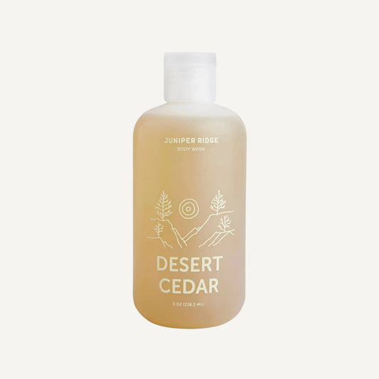 A bottle of Juniper Ridge Body Wash in the scent 'Desert Cedar', featuring a frosted translucent bottle filled with a light amber liquid and a white cap, set against a plain white background.