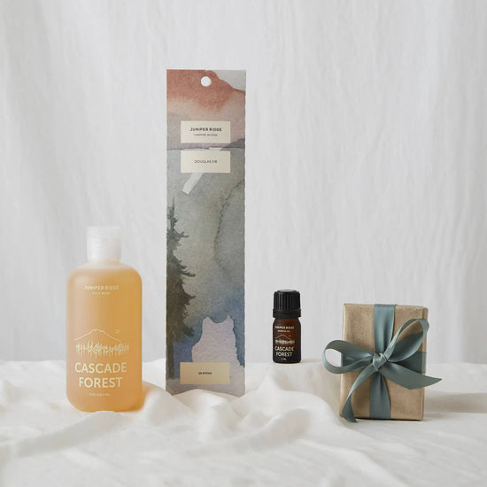 Juniper Ridge Campfire Incense in Douglas Fir gift set featuring a translucent amber bottle, a tall landscape-themed watercolor box, a small dark amber essential oil bottle, and a kraft paper gift box tied with a sage green ribbon, arranged on a white fabric surface.
