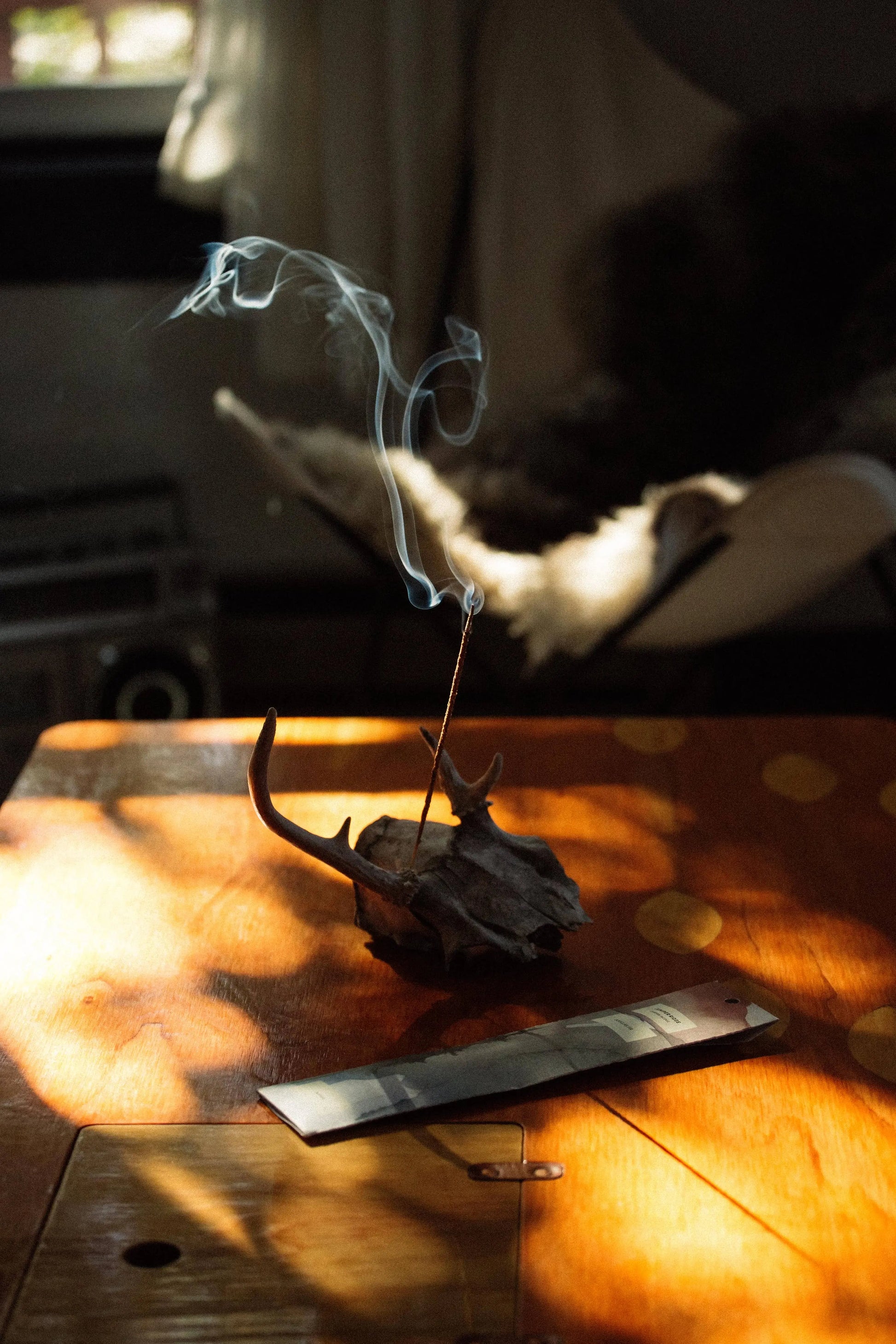 A rustic incense holder made from a deer skull with antlers sits on a wooden table in warm, dappled sunlight, with a incense stick burning and smoke curling upward. A metal tray is placed in front of the holder.