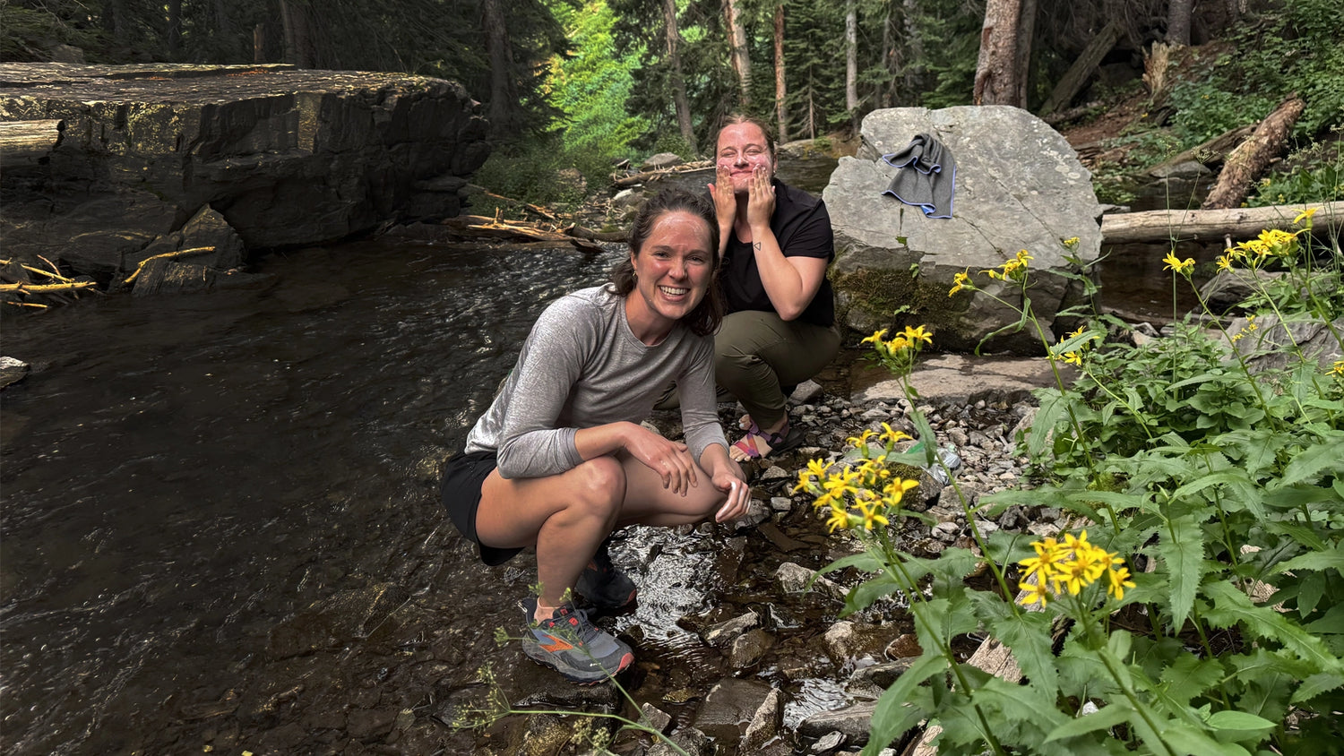 Two young ladies wash their face in a mountain creek with Simple Body Foaming Face wash while surrounded by nature.
