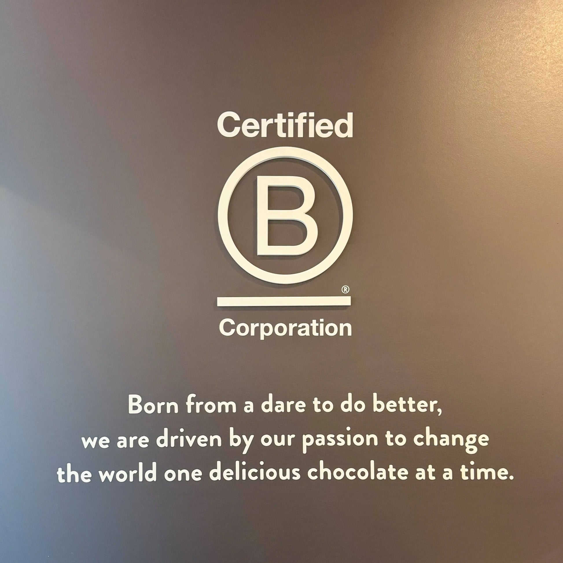 Certified B Corporation logo and text on a dark brown background, with the slogan 'Born from a dare to do better, we are driven by our passion to change the world one delicious chocolate at a time.'.