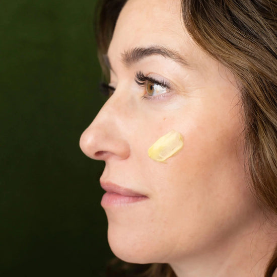 A woman showing a swatch of Simple Body Beauty Balm on her cheek with a green background