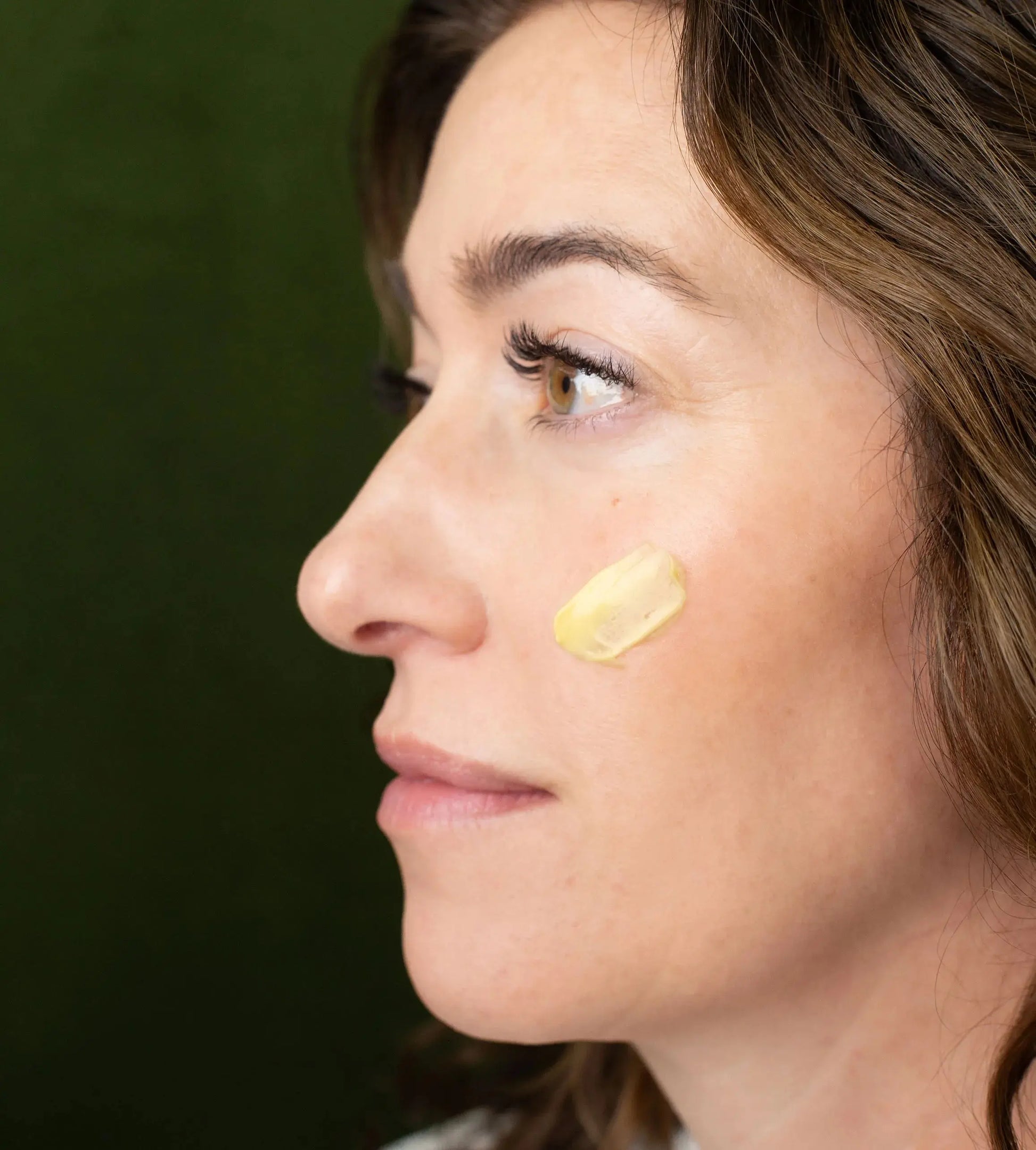 A woman showing a swatch of Simple Body Beauty Balm on her cheek with a green background