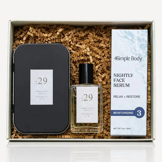 A skincare gift set in a white box filled with brown crinkle paper. The set includes a black metal tin, a clear glass bottle of No 29 Amber Wood Vanilla Perfume Oil, and a Simple Body Nightly Face Serum.