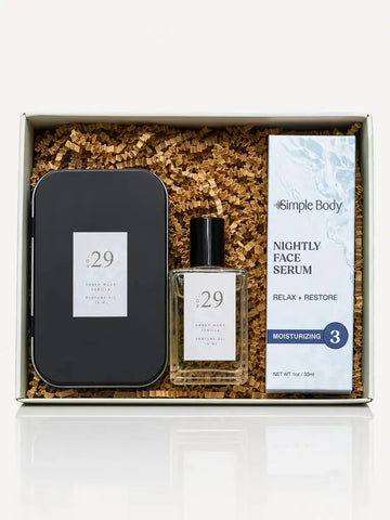 A skincare gift set in a white box filled with brown crinkle paper. The set includes a black metal tin, a clear glass bottle of No 29 Amber Wood Vanilla Perfume Oil, and a Simple Body Nightly Face Serum.