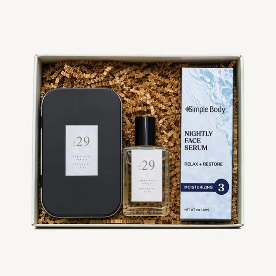 A skincare gift set in a white box filled with brown crinkle paper. The set includes a black metal tin, a clear glass bottle of No 29 Amber Wood Vanilla Perfume Oil, and a Simple Body Nightly Face Serum.