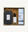 A skincare gift set in a white box filled with brown crinkle paper. The set includes a black metal tin, a clear glass bottle of No 29 Amber Wood Vanilla Perfume Oil, and a Simple Body Nightly Face Serum.