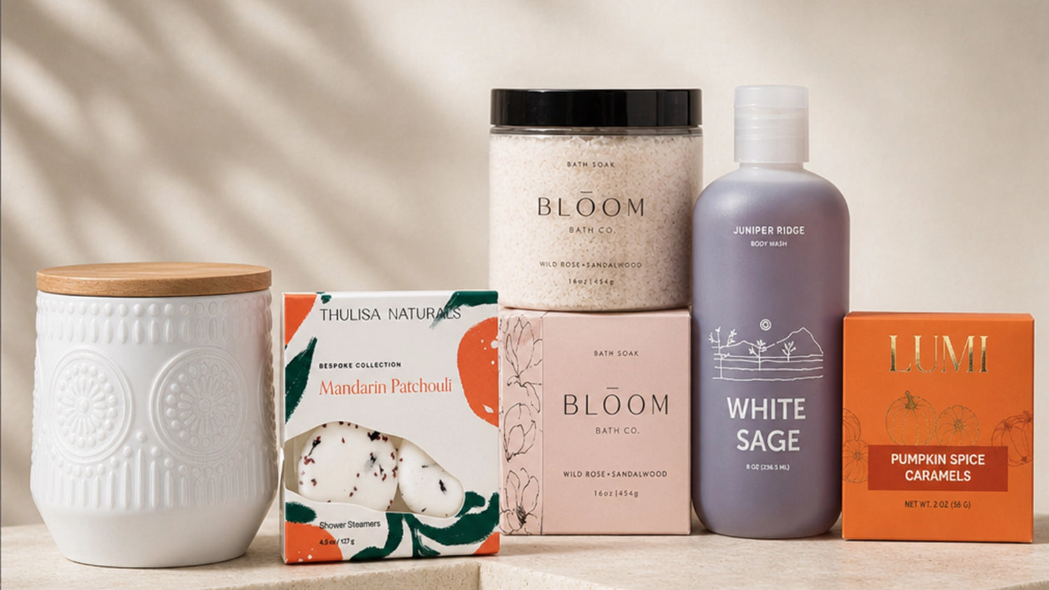 A curated collection of bath products arranged on a light wood surface against a warm beige background with soft leaf shadows. The products include a white ceramic candle jar with a bamboo lid, a jar of Bloom bath salts, a bottle of White Sage shower gel, a box of Mandarin Patches, and a box of Lumi pumpkin spice caramels.