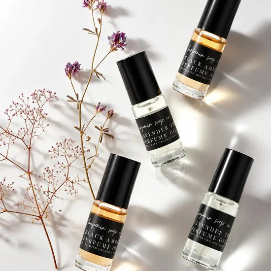 Four clear glass perfume oil bottles from Benjamin Soap Co with black caps are arranged on a white background with dried floral stems. The labels read 'Black Amber and Lavender Thyme,' and 'Perfume Oil'.