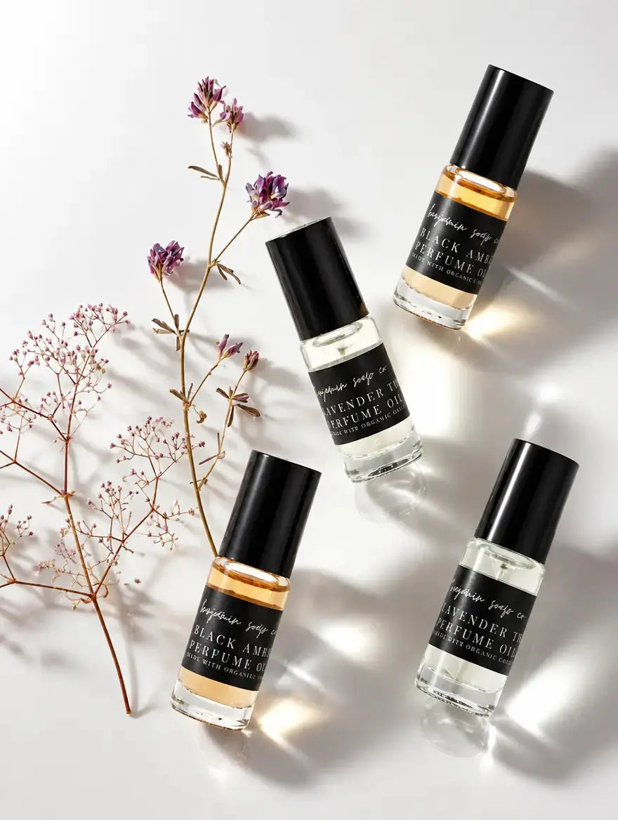 Four clear glass perfume oil bottles from Benjamin Soap Co with black caps are arranged on a white background with dried floral stems. The labels read 'Black Amber and Lavender Thyme,' and 'Perfume Oil'.