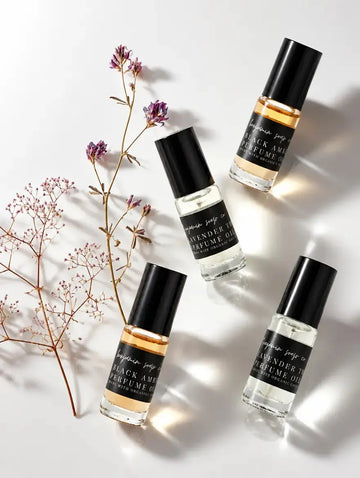 Four clear glass perfume oil bottles from Benjamin Soap Co with black caps are arranged on a white background with dried floral stems. The labels read 'Black Amber and Lavender Thyme,' and 'Perfume Oil'.