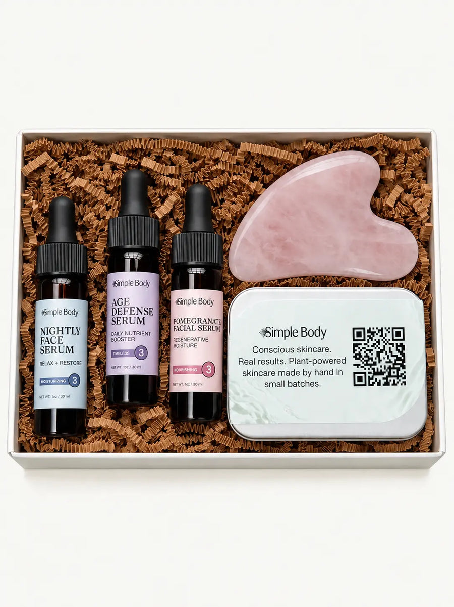A skincare gift set from Simple Body arranged in a white box filled with brown crinkle paper. The set includes three small dropper bottles labeled Nightly Face Serum, Age Defense Serum, and Pomegranate Facial Serum, a mint green gua sha face scraper, and a white branded tin.