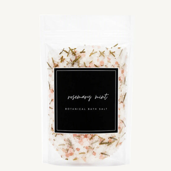 A clear stand-up pouch of rosemary mint botanical bath salt, showing the white bath salts mixed with pink crystals and green herbs through a black label on a light gray background.