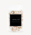 A clear stand-up pouch of rosemary mint botanical bath salt, showing the white bath salts mixed with pink crystals and green herbs through a black label on a light gray background.