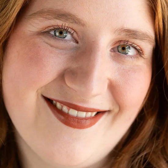 Close-up of a person with red hair and green eyes, wearing orange-brown warm lipstick by Pure Anada.