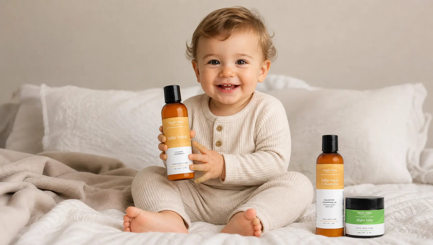 A smiling toddler sits on a bed holding an amber bottle of Simple Mama Baby Lotion, with two more Simple Mama products standing beside them. The room has soft neutral bedding and pillows in the background.