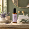A bottle of Simple Body Age Defying Tonic with a lavender label and black cap, standing on a stone countertop in a bathroom setting with lavender flowers, white towels, and a mirror in the background.