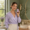 A woman in a lavender wrap blouse and cream trousers holds up a small Simple Body Age Defying Botanical Skin Tonic bottle with a black cap and a light purple label. She is standing in a bathroom with a green wall, a round mirror, candles, and a sink in the background.
